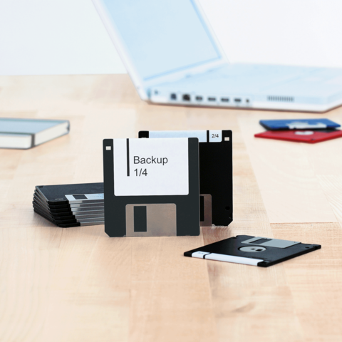 Mehrere Disketten liegen auf einem Tisch, eine mit der Aufschrift "Backup 1/4". Im Hintergrund ist ein Laptop sichtbar.